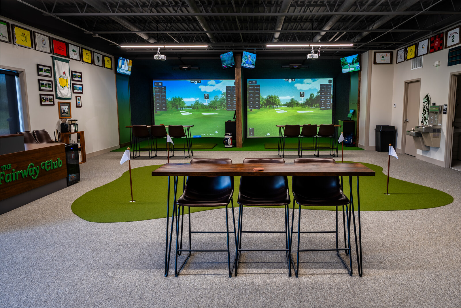 The interior of Fairway Club in Lee's Summit, Missouri &mdash; two simulator bays, a central putting green with a high-top table, and walls lined with framed major championship pin flags from the Masters, U.S. Open, PGA Championship, and Pinehurst No. 2