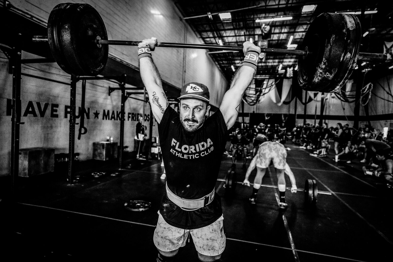 Zac Ansaldo locking out a barbell overhead in a Florida Athletic Club t-shirt and Kansas City Chiefs hat, in a black-and-white photo with 'HAVE FUN, MAKE FRIENDS' visible on the gym wall behind him