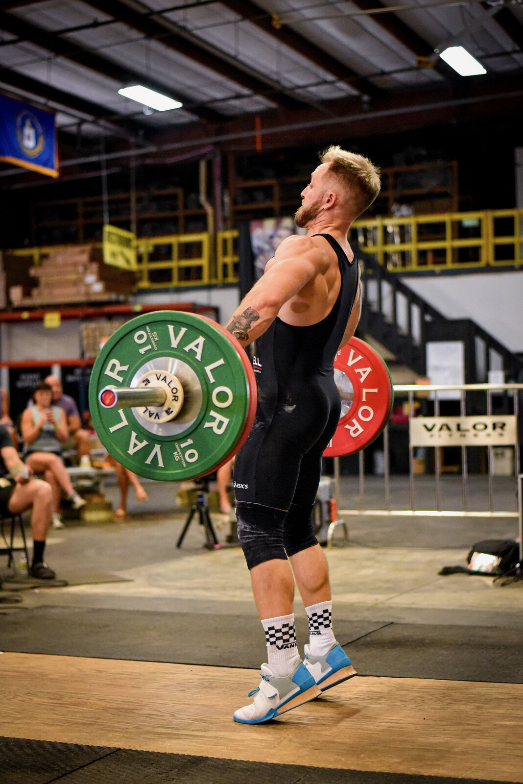 Zac Ansaldo pulling a barbell during an Olympic weightlifting competition, wearing a black singlet with green and red Valor competition plates loaded on the bar